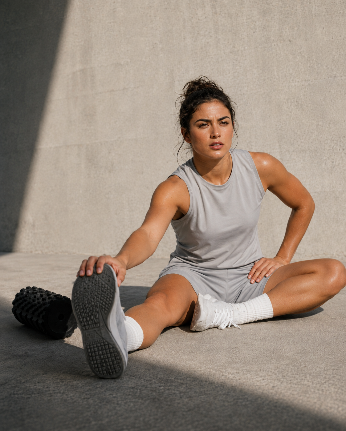 A woman in a light gray athletic tank top and matching shorts sits on a concrete surface stretching, with one foot raised. She is wearing white socks and a black textured athletic shoe, with another shoe placed beside her.