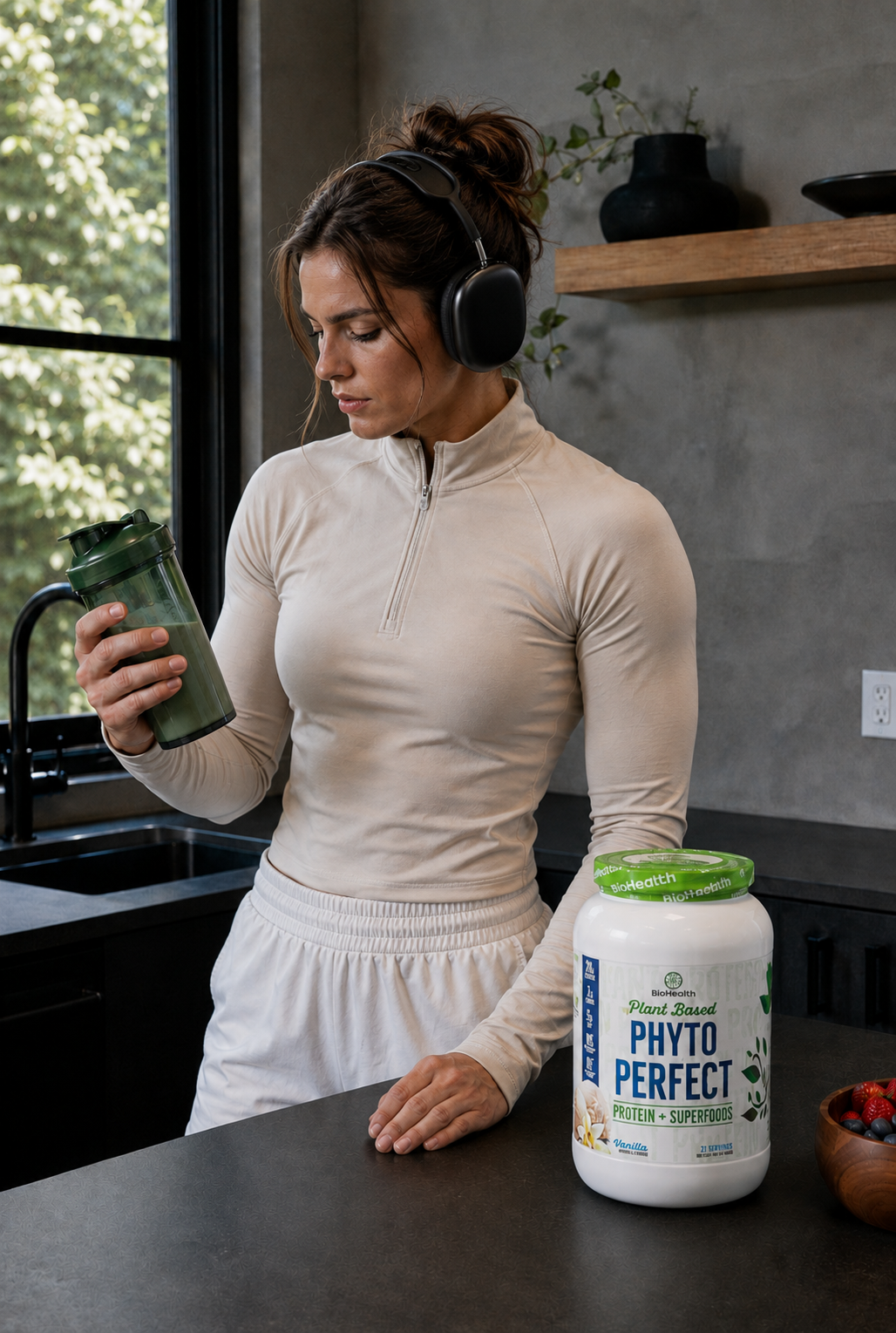 A woman in a beige athletic quarter-zip top and white sweatpants stands at a kitchen counter wearing headphones and holding a green shaker bottle. A large tub of Plant Based Phyto Perfect + Green Superfood powder sits on the counter.