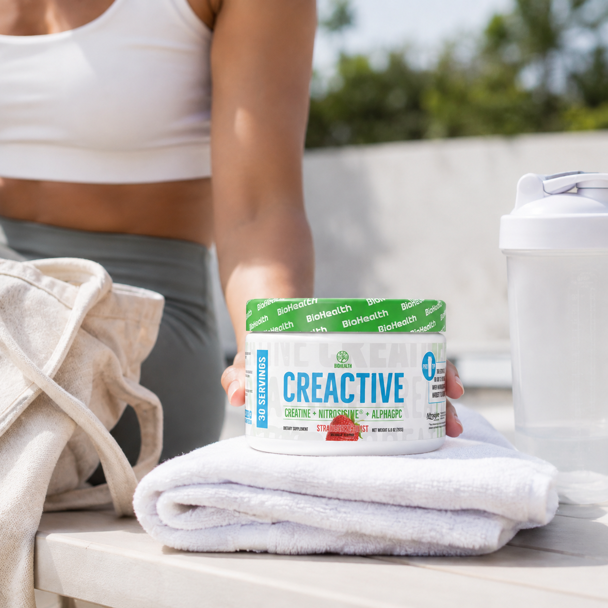 A person in a white sports bra and gray leggings holds a white tub of CREATIVE dietary supplement powder on a white table, with a beige tote bag, white towel, and white shaker bottle nearby in an outdoor setting.