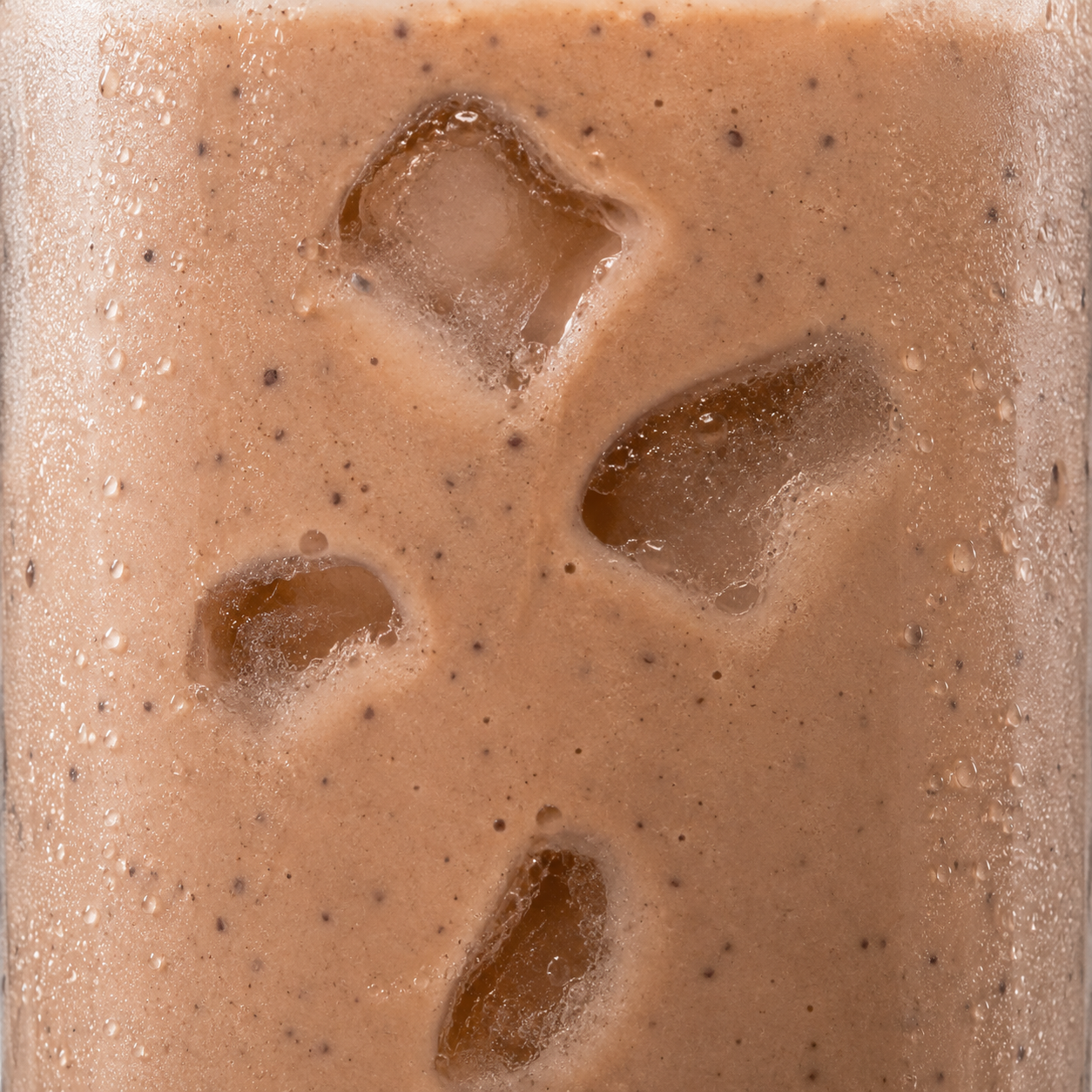 A close-up of a thick, creamy light brown beverage with visible ice cubes and tiny dark specks, filling the frame.