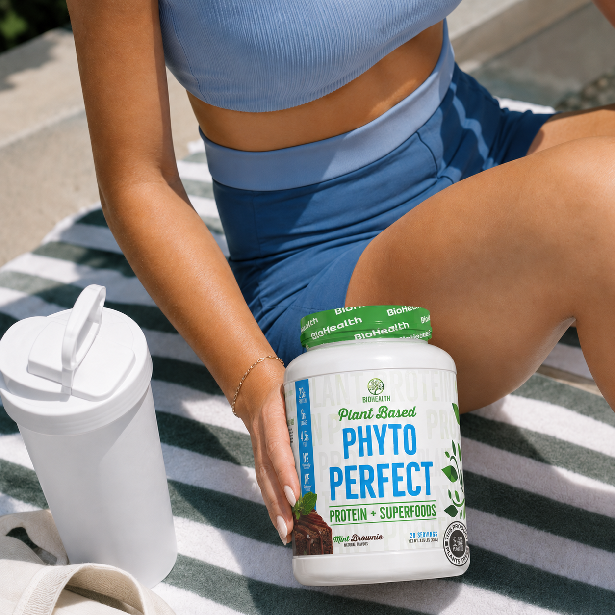 A woman in blue athletic wear sits on a striped blanket outdoors, holding a tub of PlantBased Phyto Perfect protein powder. A white shaker bottle is also visible on the blanket next to the tub.